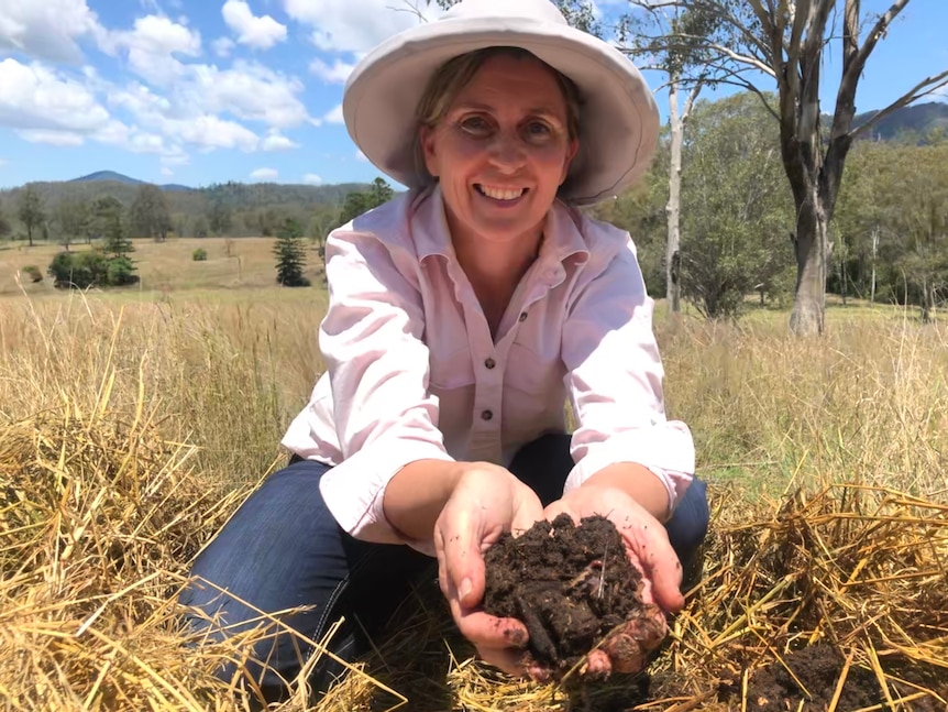 A lady crouches down holding out a handful of soil and worms towards the camera.