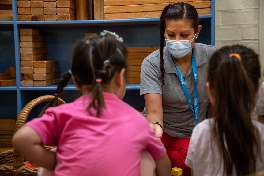 A childcare worker wearing a surgical mask plays with two children.