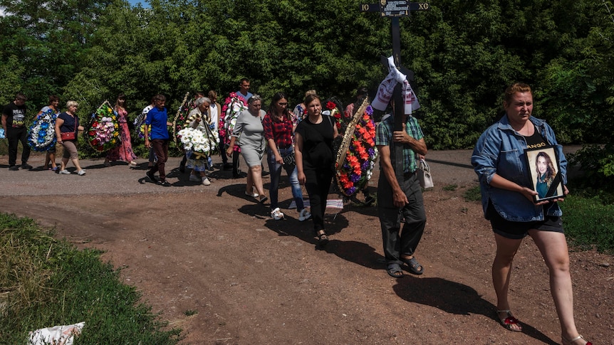 People carrying flowers and tributes walk in a straight line along a dirt road. The woman at the front is holding a photo.