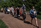 People carrying flowers and tributes walk in a straight line along a dirt road. The woman at the front is holding a photo.