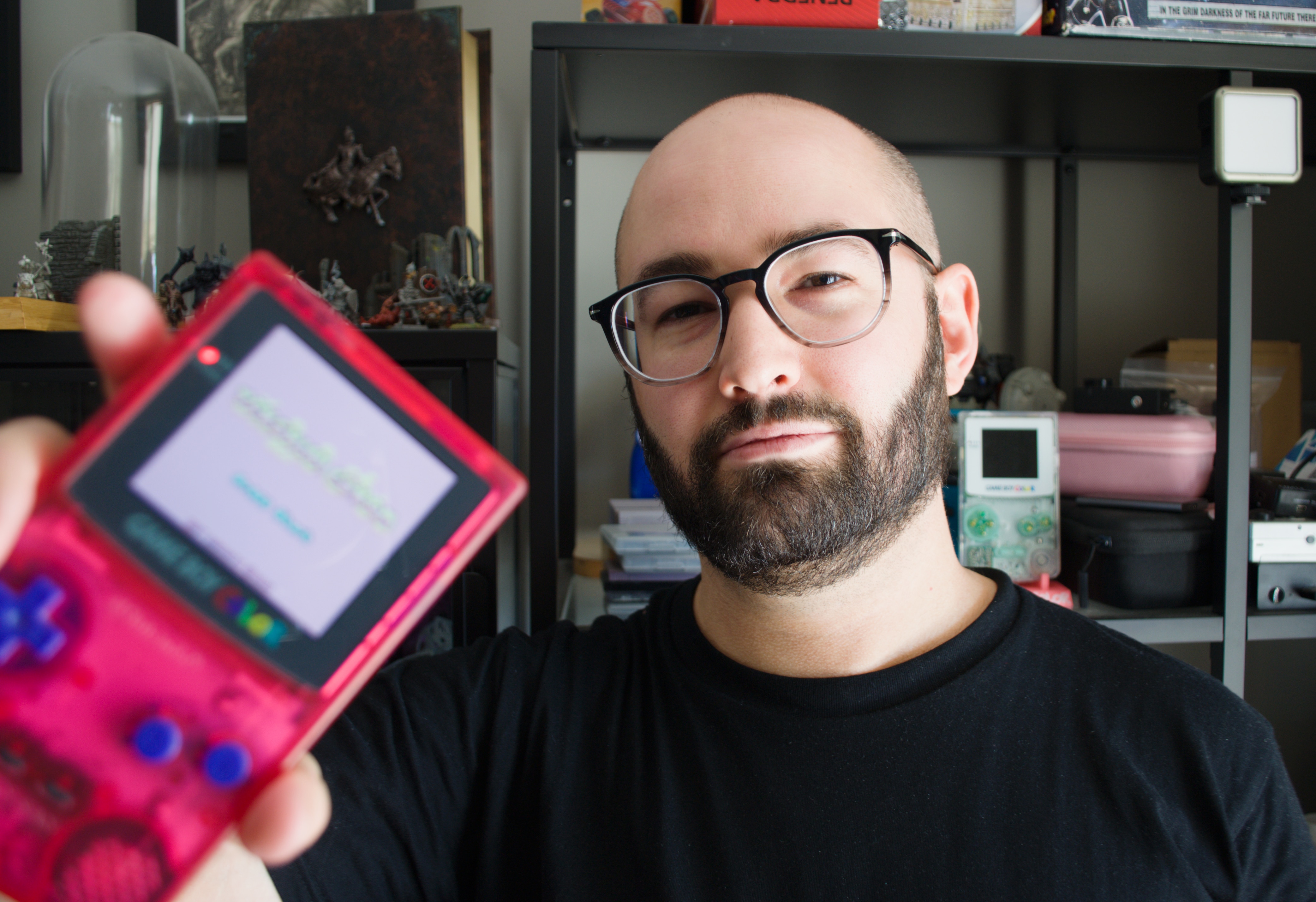 A man with glasses and a beard holds a gameboy towards the camera which is pink with a backlit screen