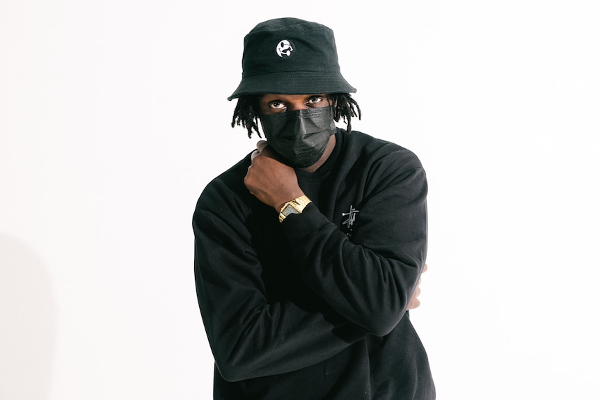 A man with dreadlocks wearing a black bucket hat and black face-mask looks at the camera