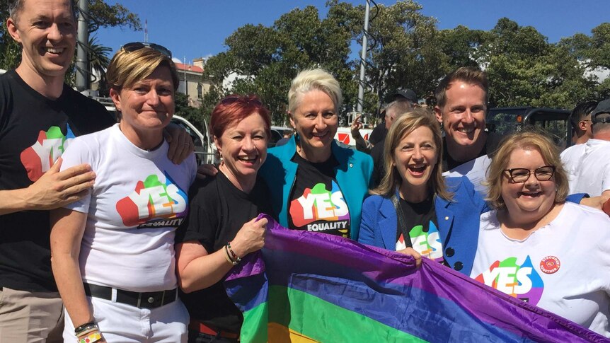 A group of men and women wearing 'yes' shirts stand with a rainbow flag with big smiles