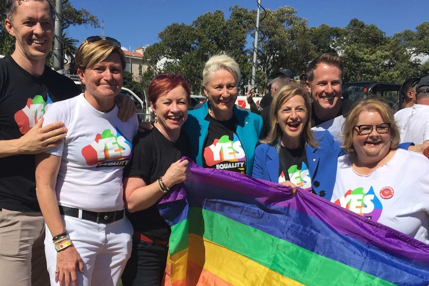 A group of men and women wearing 'yes' shirts stand with a rainbow flag with big smiles