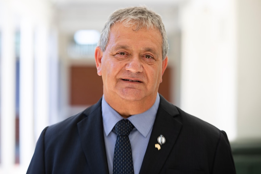 A man in a shirt, tie and jacket with pins on it smiles at the camera.