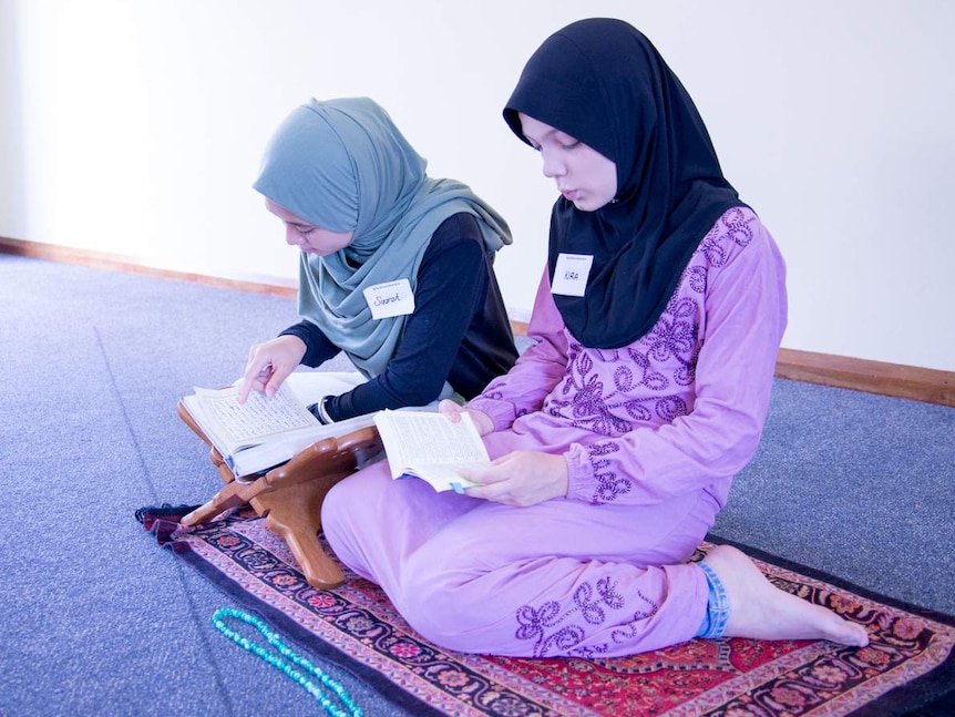 Two Islamic women dressed in hijabs kneel on a rug while reading passages from the Koran