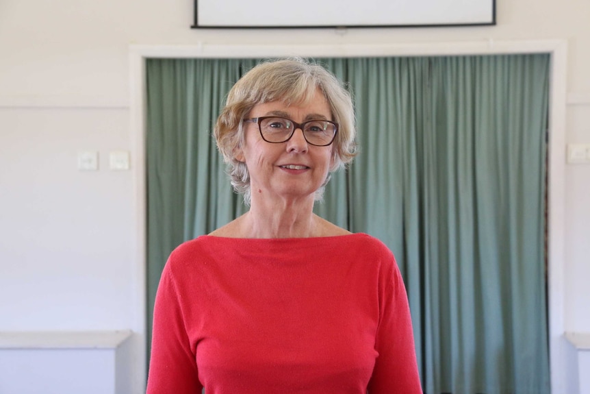 A well-groomed elderly woman in glasses and red blouse stands in front of green curtain in room.