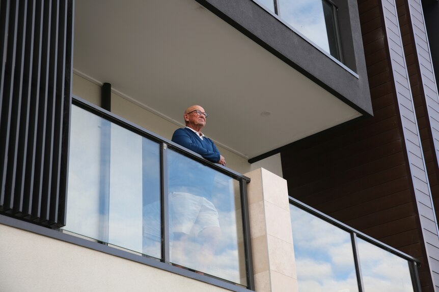 A man wearing a blue jumper looks out over a glass balcony, viewed from below.