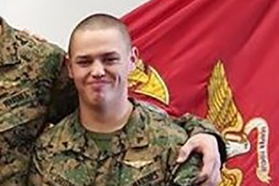 A young US marine stands in front of a flag.