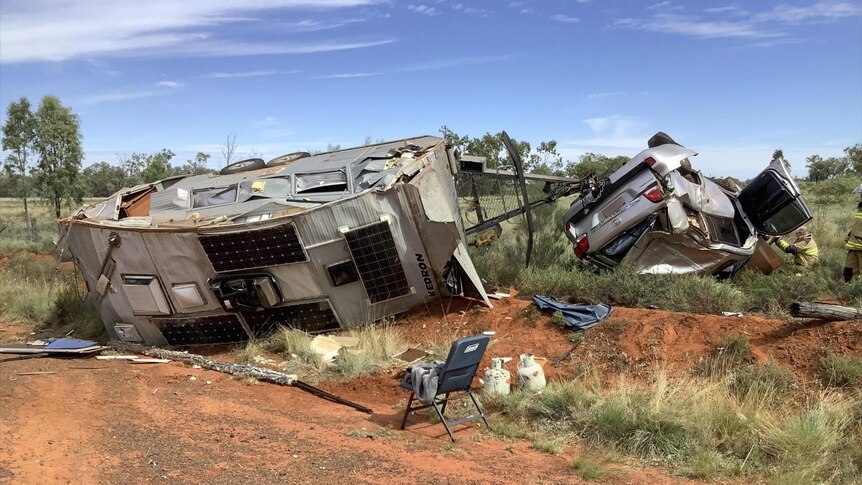 A crashed, upside down 4wd and caravan on it's side of a red dirt highway. 