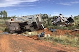 A crashed, upside down 4wd and caravan on it's side of a red dirt highway. 
