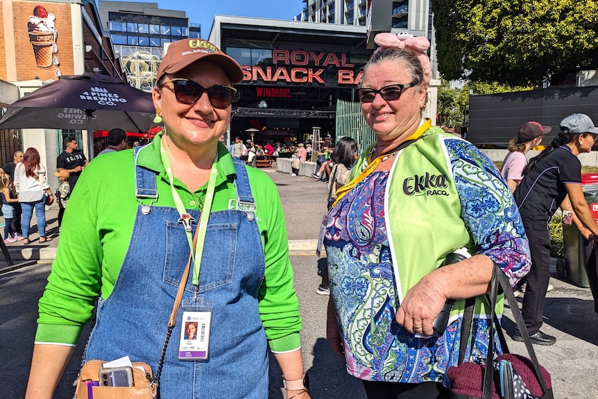 Two woman in bright green shirts smile in front of a carnival food stall. Their shirts say "Ekka 2023".