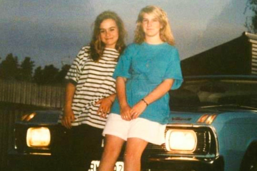 An old photo of two teenage girls leaning against a car giggling.