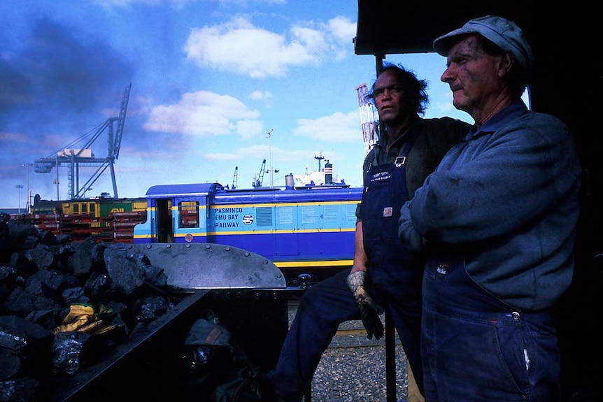Two sooty faced men in train driving overalls atop a steam train loco.