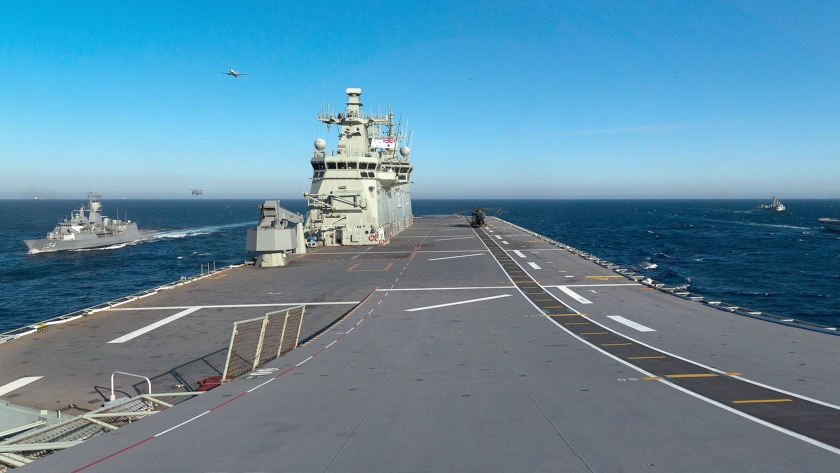 Three naval ships and military planes travel in formation with a carrier on the ocean.