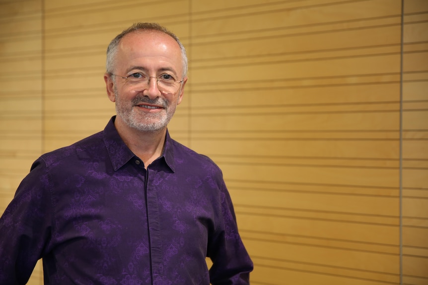 Andrew Denton - smiling, in blue shirt and with short, grey facial hair - stands in front of a wooden wall.