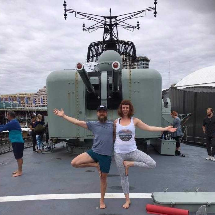 A man and a woman both stand on one leg in a yoga pose on board a ship with a large gun visible in the background
