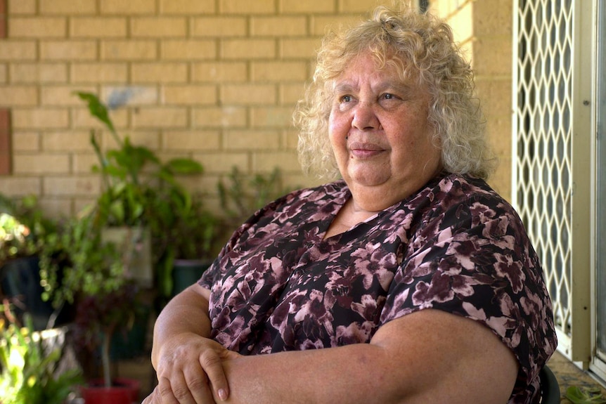 A woman sits on a balcony of a brick home, looking forward with a neutral expression, her hand on her lap.