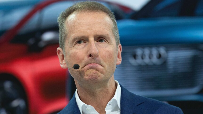 Close-up shot of Herbert Diess as he stand in front of a car with a microphone headset on.