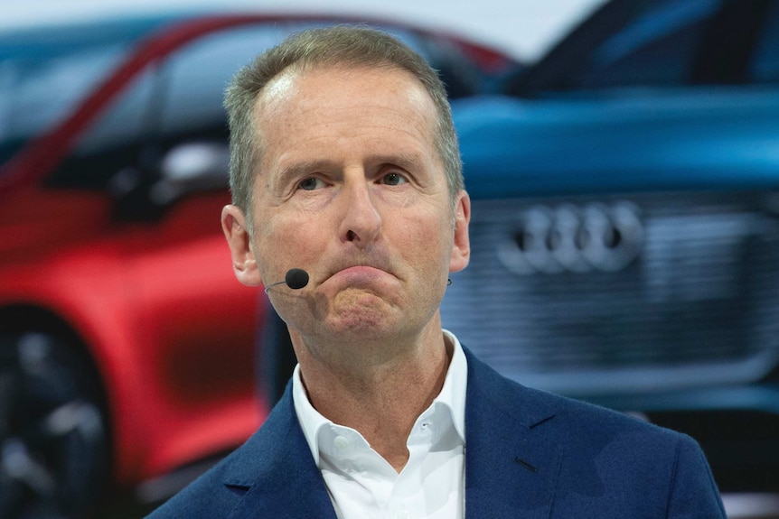 Close-up shot of Herbert Diess as he stand in front of a car with a microphone headset on.