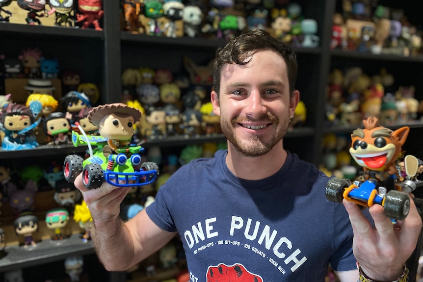 smiling man holds up a toy story and crash bandicoot pop vinyl figurine 