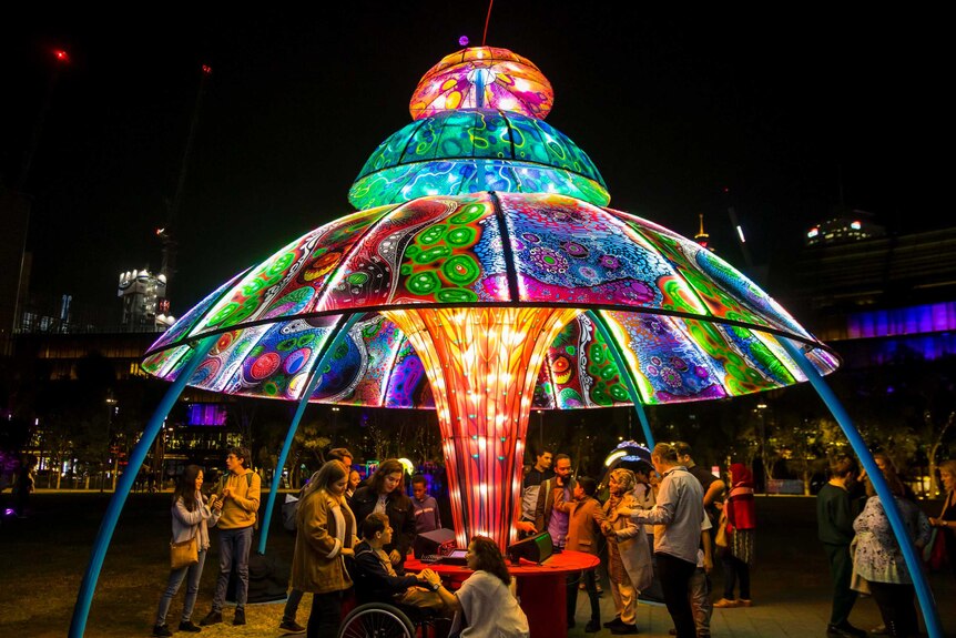 People, including a man in a wheelchair, gather underneath a large dome of colourful lights.