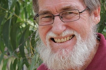 A man with glasses and a beard smiling behind a green leafy tree
