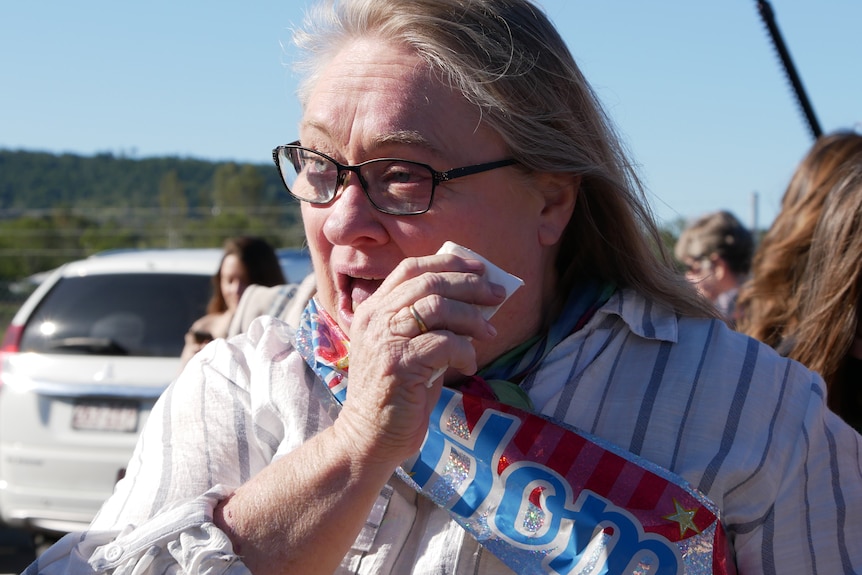 A woman with long gray hair wipes away tears as she stands outside an airport.