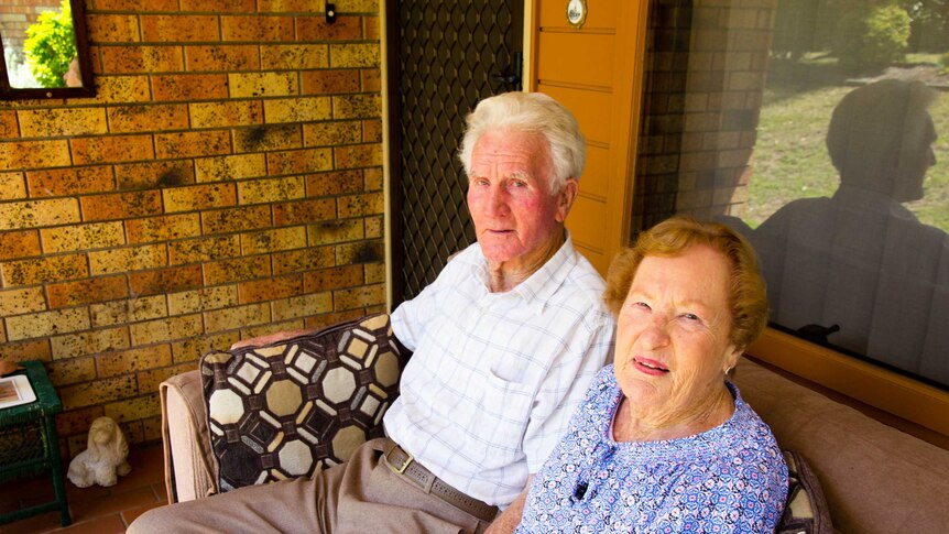 An elderly couple sitting on a couch outside a brick house holding hands.