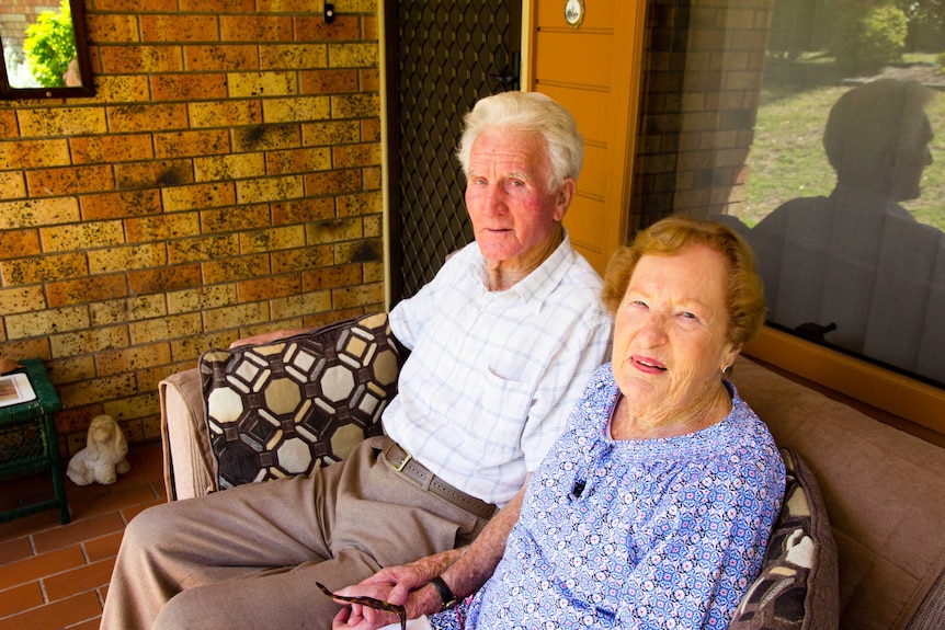 An elderly couple sitting on a couch outside a brick house holding hands.
