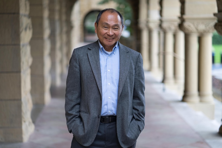 A man in a gray suit and blue shirt poses for a photo in a breezeway flanked by sandstone columns