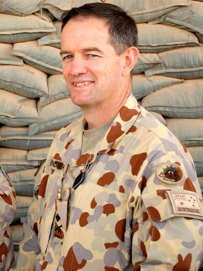 General Mike Hindmarsh, wearing camouflage Army clothing, smiles at the camera in front of a stack of sandbags.