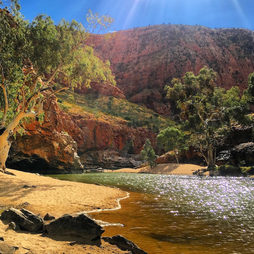 Blue sky, deep orange rock, white trunked gum trees and a water hole.