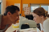 A middle-aged man leans over a donut shop counter to speak to a bemused-looking young woman 