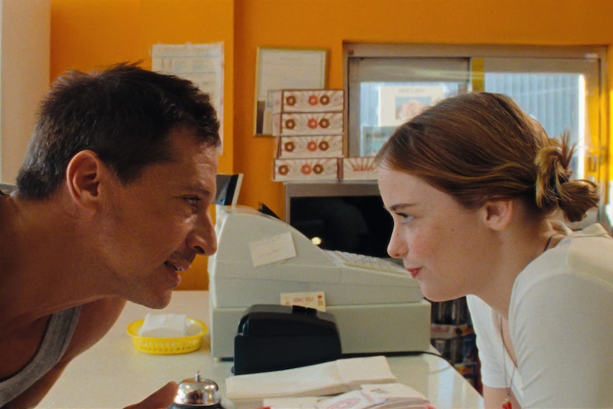 A middle-aged man leans over a donut shop counter to speak to a bemused-looking young woman 