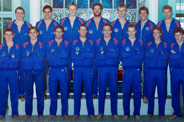 A group of young men dressed in blue tracksuits stand in two rows with their arms folded and their coach at the end of front row