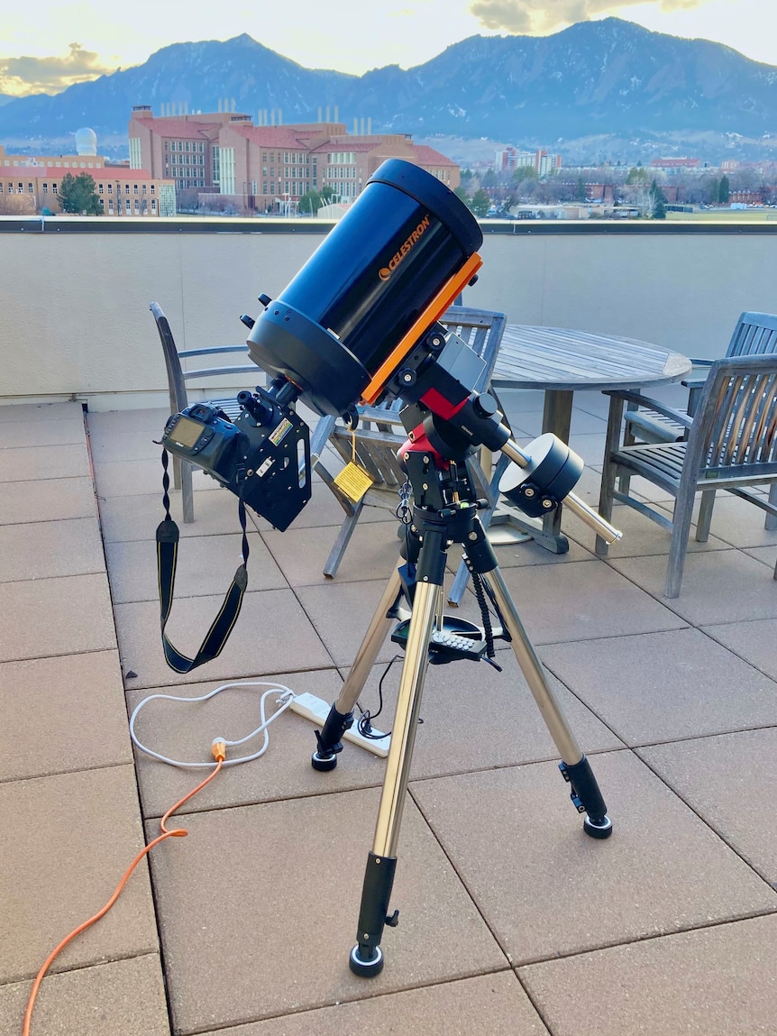 A telescope and tripod on the balcony of a mid-rise building in a town with mountains in the background.