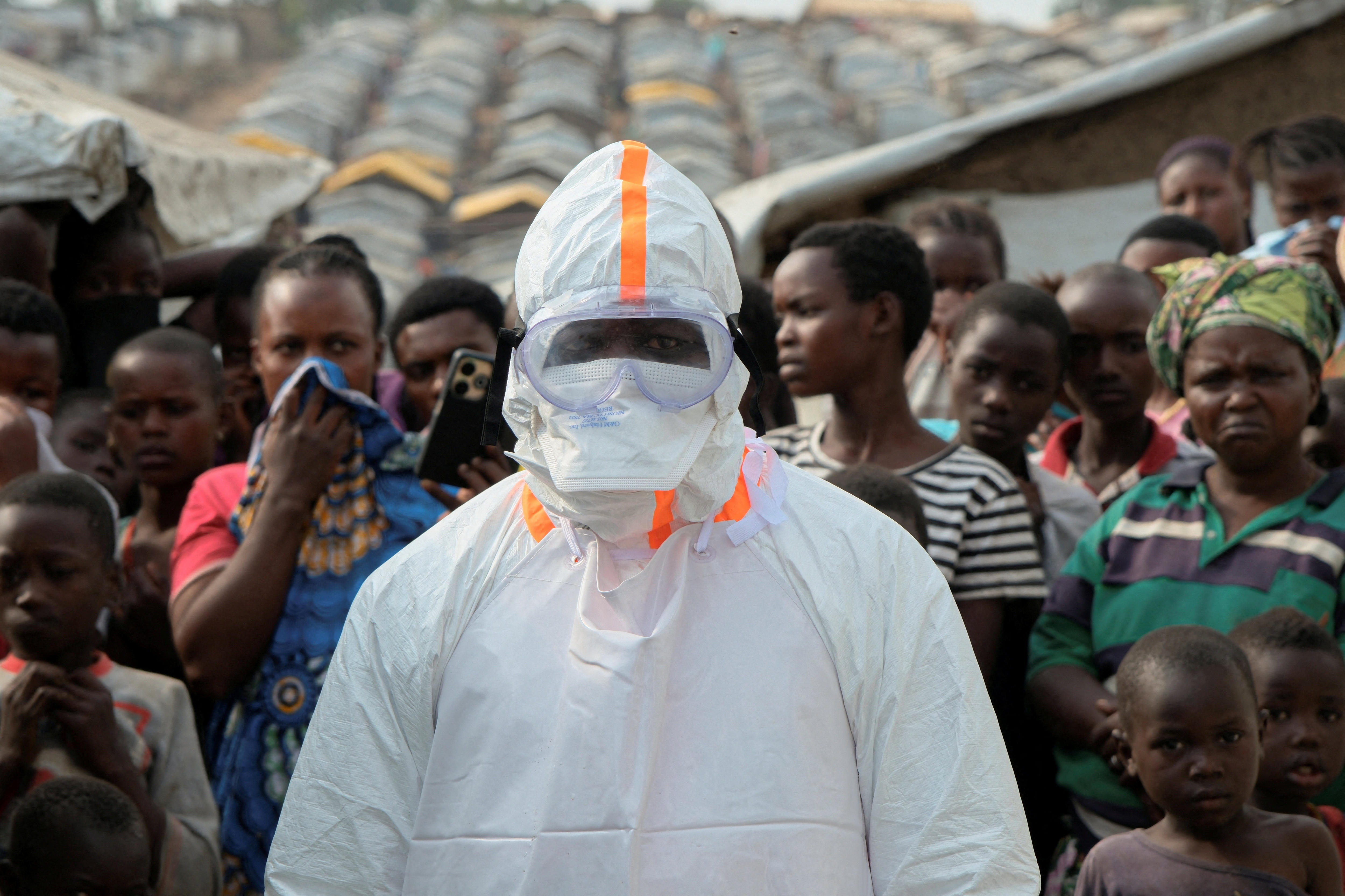 A health worker in personal protective equipment (PPE) stands near displaced people in the DRC