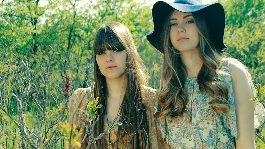 Two members of First Aid Kit stand in a forest. One wears a wide-brim hat.
