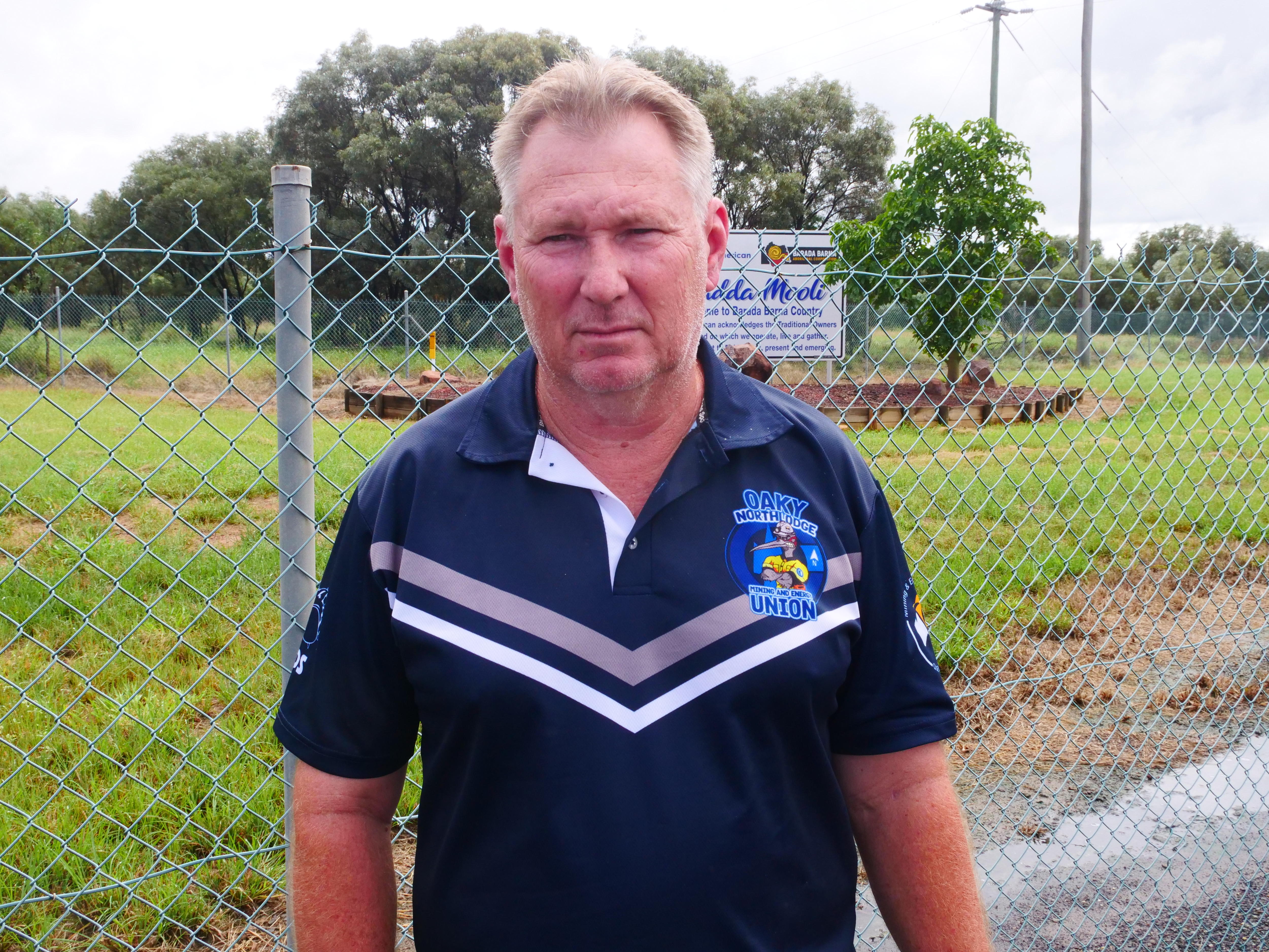 Un hombre con una camisa a rayas, mirando directamente a la cámara, con una cerca y una hierba detrás de él. 