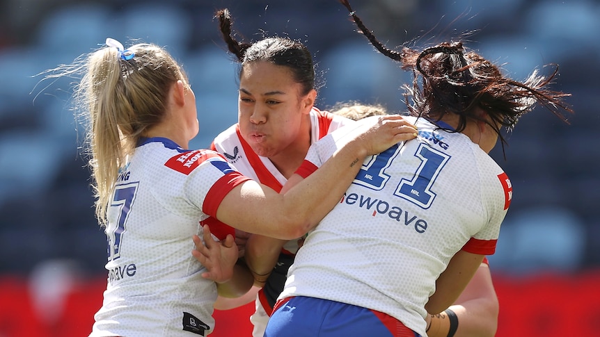 A Sydney Roosters NRLW player holds the ball as she is tackled by two Newcastle opponents.