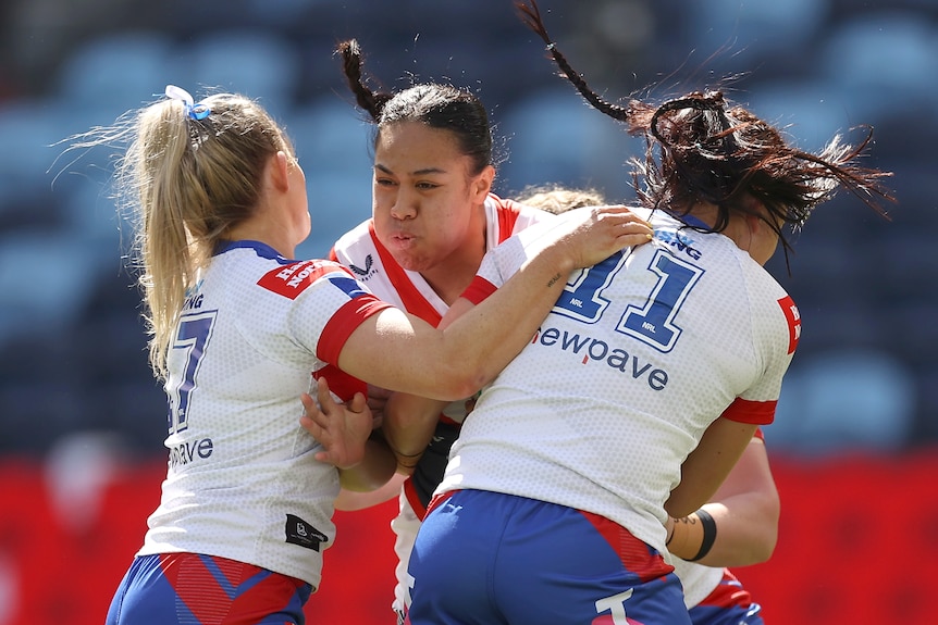 A Sydney Roosters NRLW player holds the ball as she is tackled by two Newcastle opponents.
