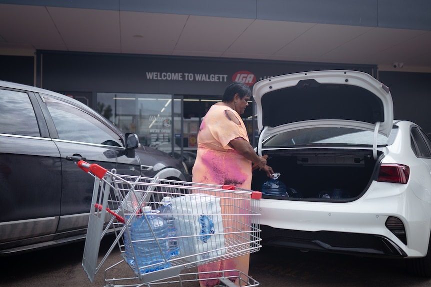 Indigenous woman in orange and pink dress loads up water bottles from a trolley into the back of her white car. 
