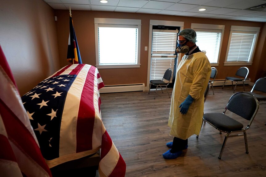 A man wearing full personal protection equipment pays respect to a coffin clad in the US flag.
