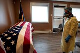 A man wearing full personal protection equipment pays respect to a coffin clad in the US flag.