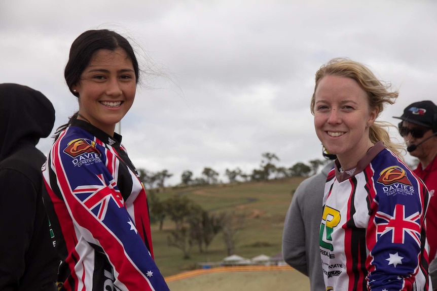 Two young female BMX riders in their gear without helmets look at the camera