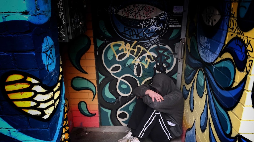 A homeless man in Hobart sits in a brightly graffitied doorway with his head in his hands.