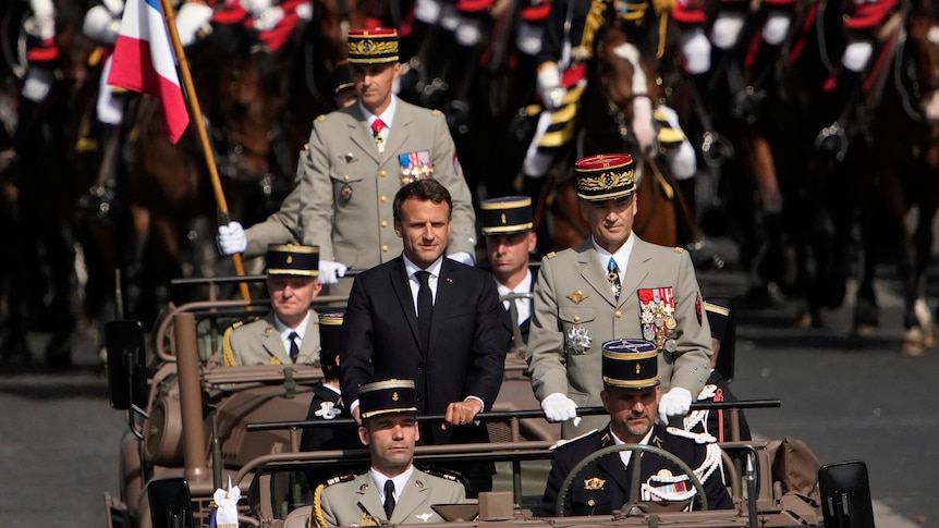 Dressed in a black suit the French President stands in the middle of an open top car surrounded by five other men. 