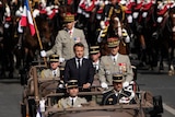 Dressed in a black suit the French President stands in the middle of an open top car surrounded by five other men. 