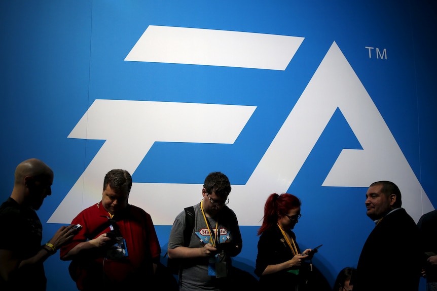 A group of men and a woman stand in front of a large EA logo looking down at their mobile phones.